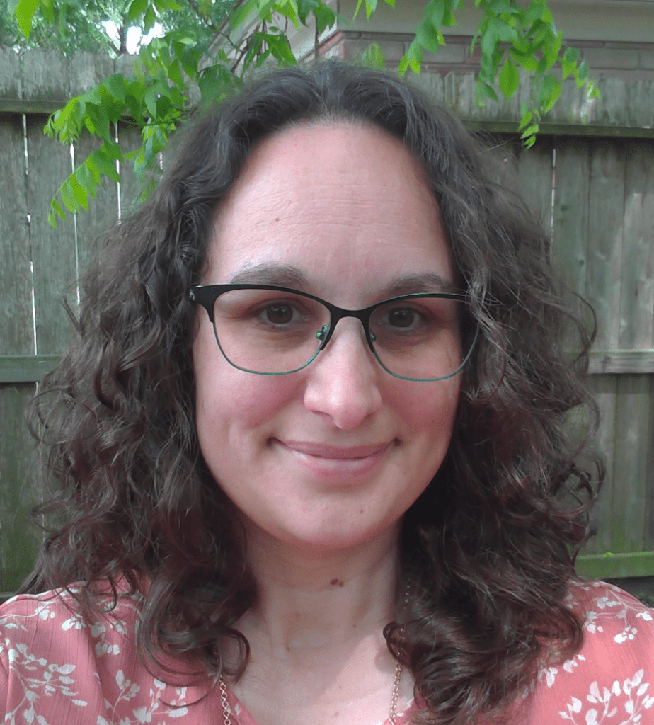 A femme person with brown curly hair and glasses is smiling at the camera. The picture is framed from the shoulders up.