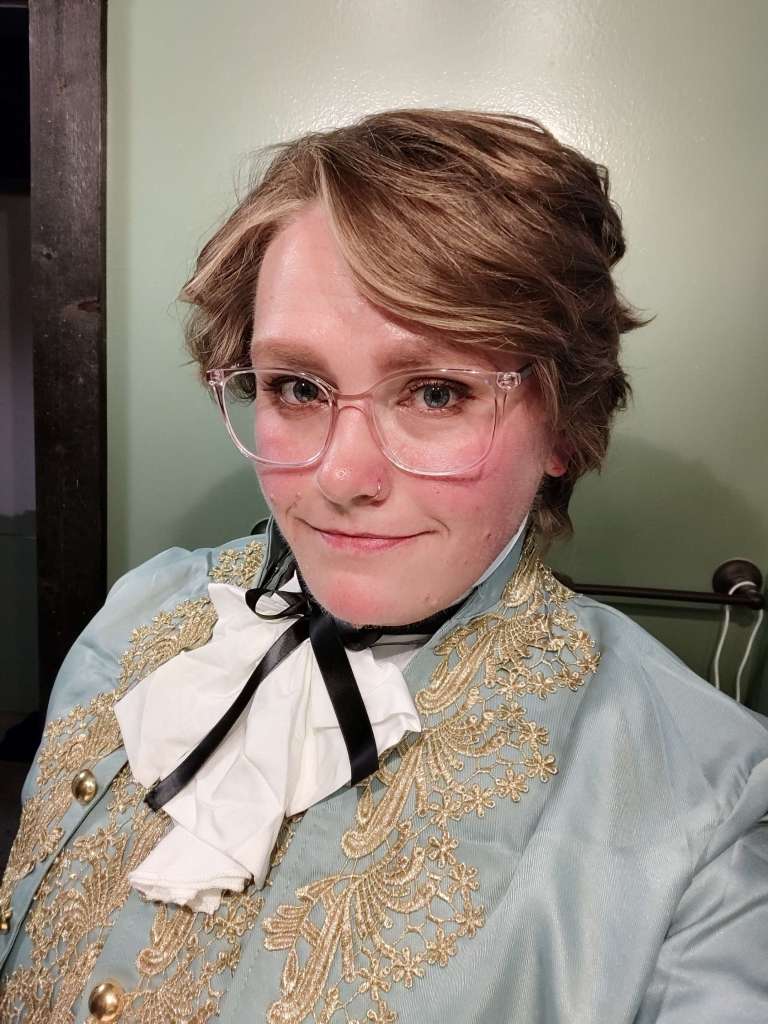 A femme person with blonde hair, glasses, and blue eyes is dressed like Stede from Our Flag Means Death.