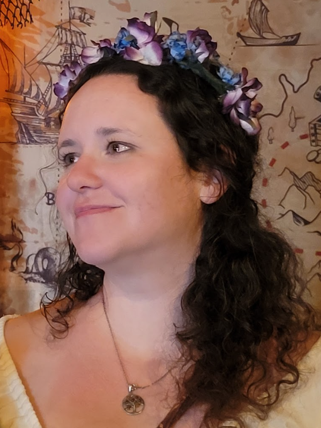 A femme person with a very cute flower crown and dark brown curly hair is smiling just off camera